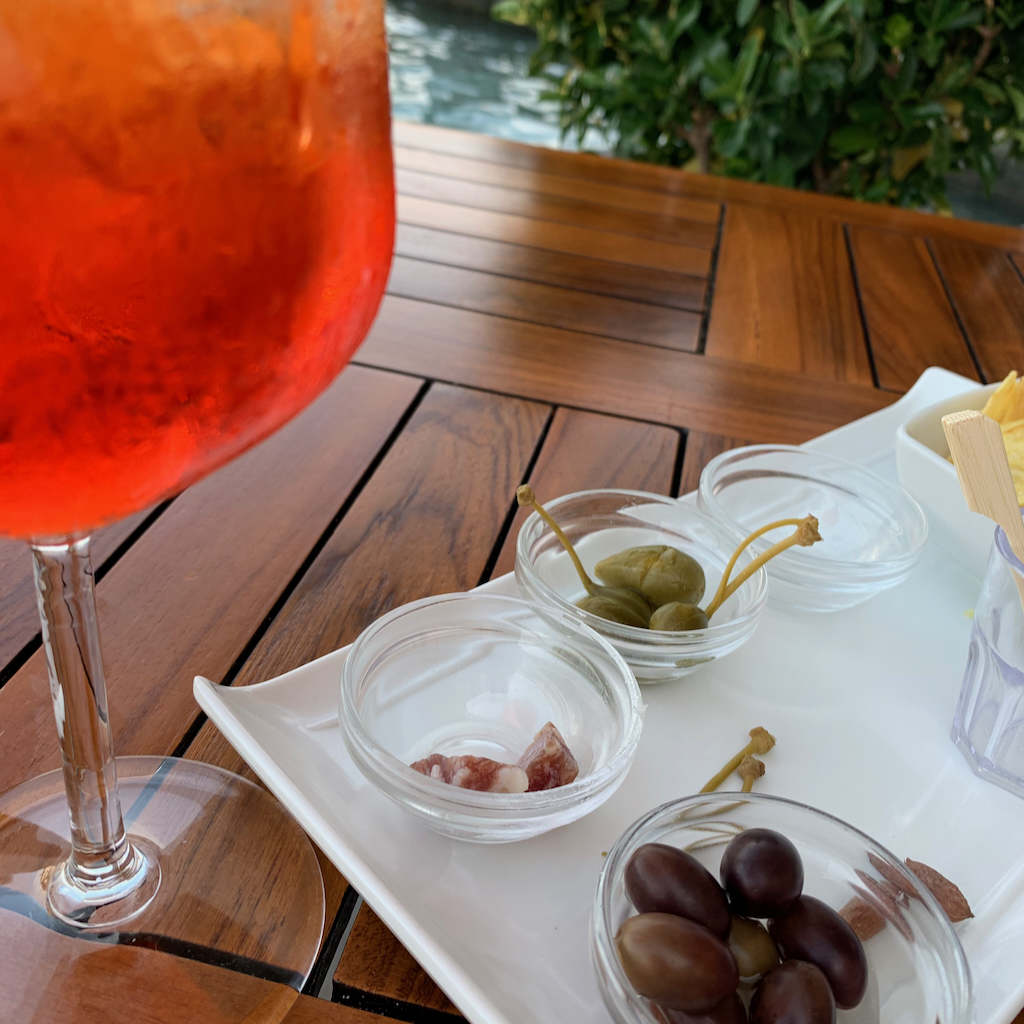 Ett immigt glas aperol spritz och aperitivo bestående av oliver, kapris, salami och chips