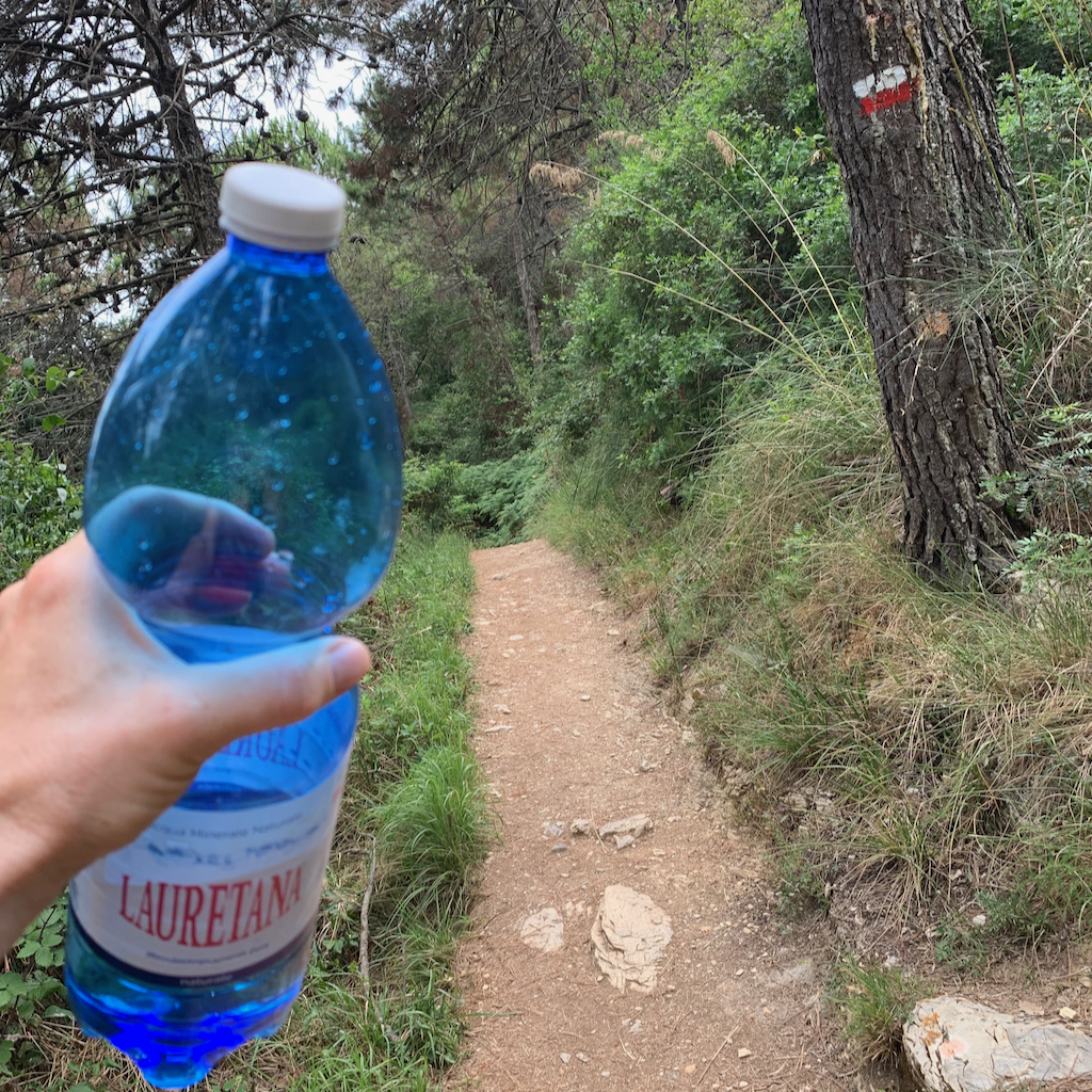 En hand med en blå 1,5-liters vattenflaska med en vandringsled i bakgrunden i Porto Venere i Cinque Terre Italien