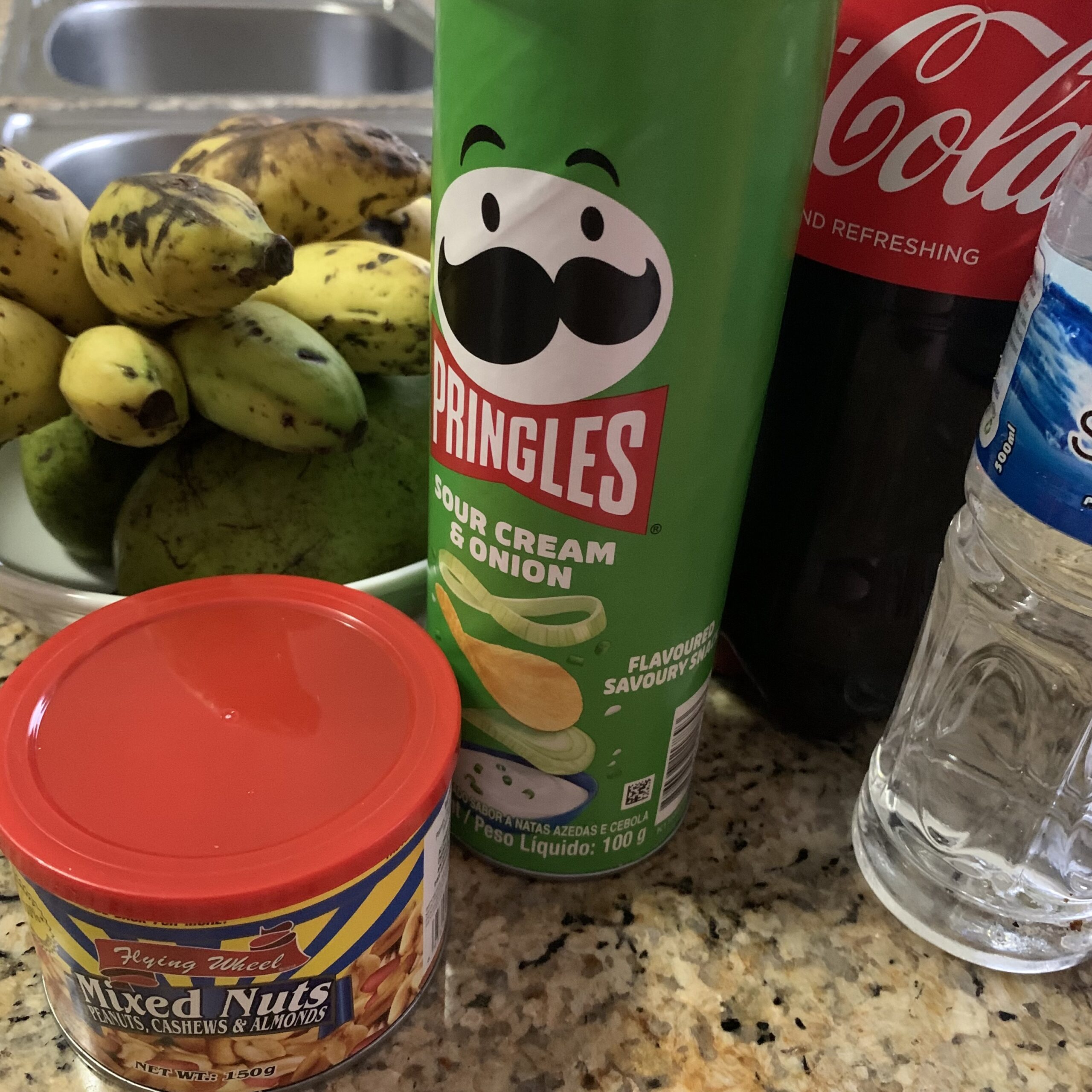 Bananer och mango i en skål bredvid en flaska cola, vatten och en burk nötter.