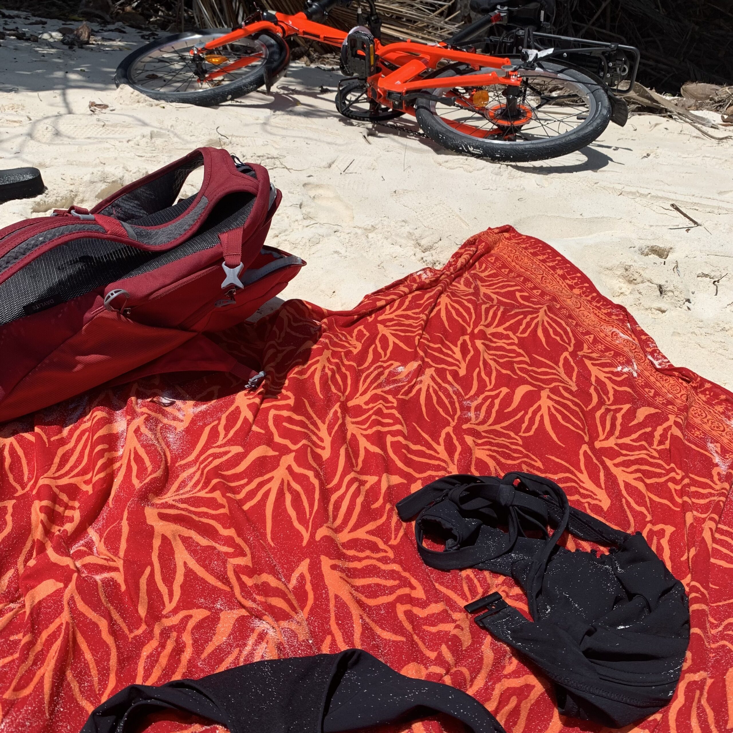 En strandsfilt med bikini och ryggsäck, samt en orange cykel i bakgrunden på en strand på Seychellerna