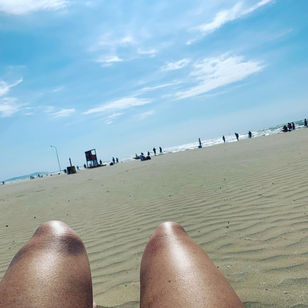 Ett par bara knän i förgrunden och en lång strand i Rimini i bakgrunden