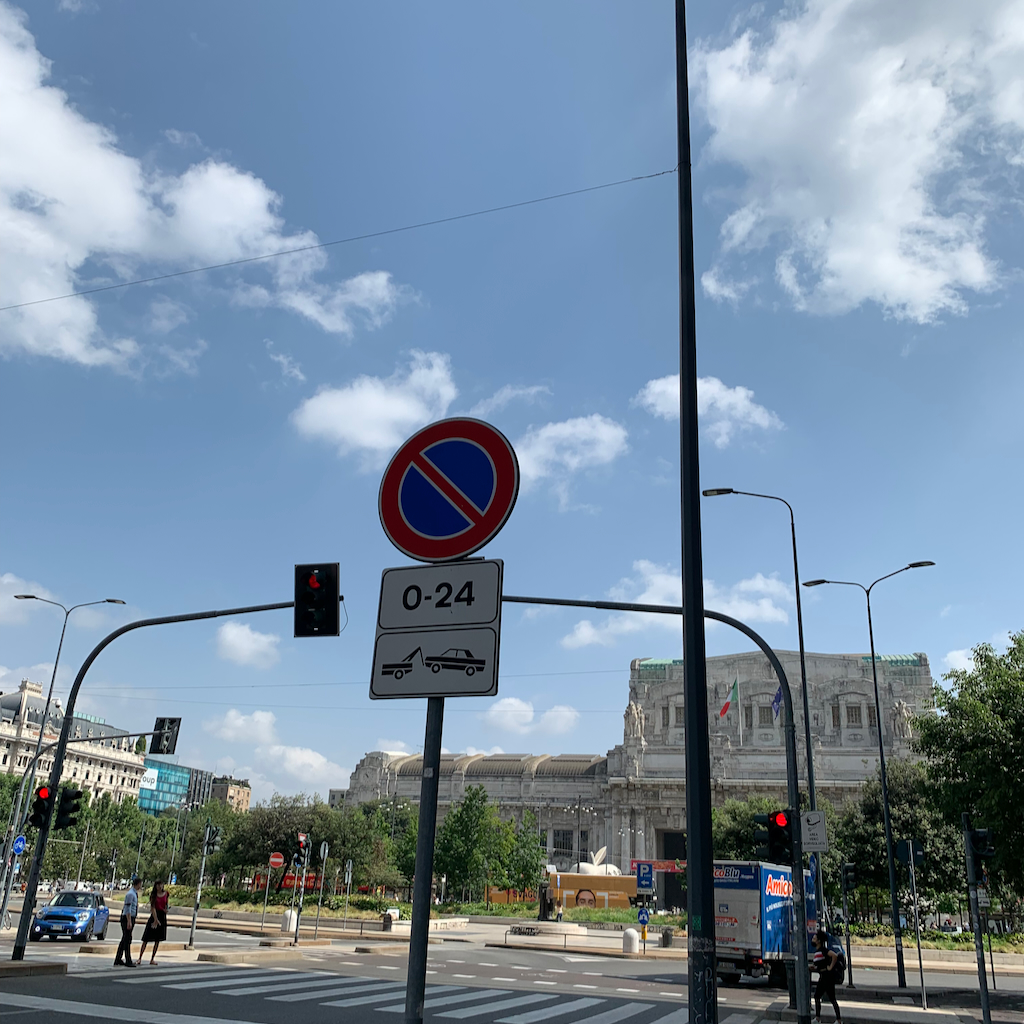 En gatukorsning med trafikljus och den massiva och pampiga Milano Termini från utsidan.