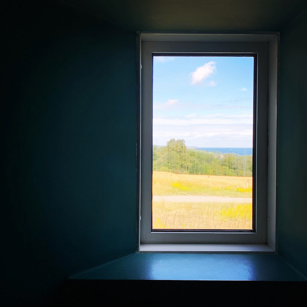 Utsikt över en sommaräng, lövskog, blått hav och himmel från ett av fönstrena på Bornholm Kunstmuseum i Danmark