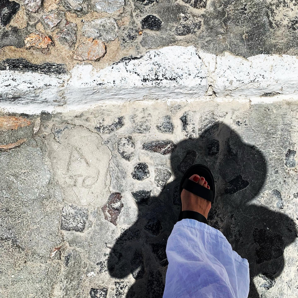 En skugga av en solhatt och fot med sandal och vita linnebyxor på vandringsleden mellan Fira och Oia på Santorini i Grekland
