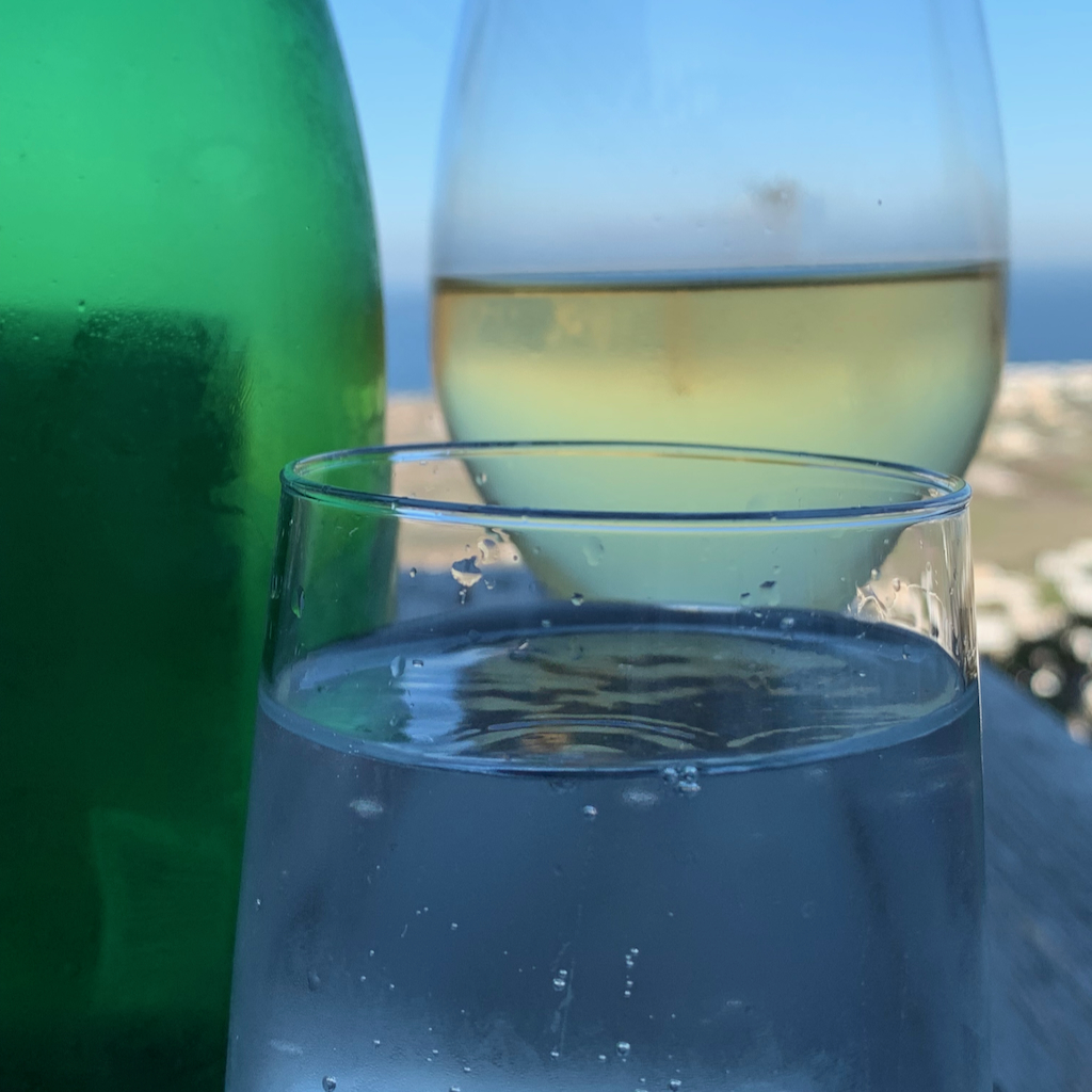 Ett glas vitt vin och en glaska vatten med havet och Kamari Beach på Santorini i Grekland i bakgrunden