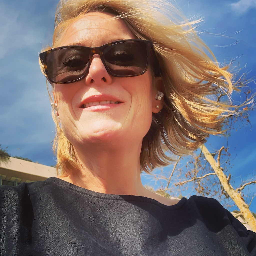 En blond kvinna med med solglasögon och vind i håret mot en blå himmel.